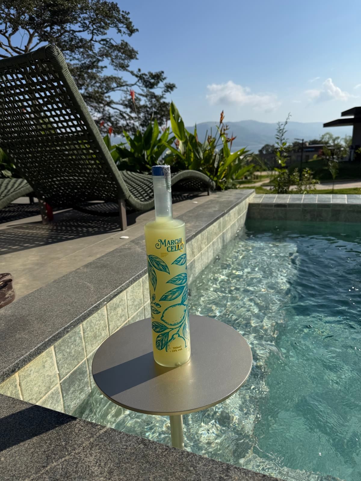 Garrafa de Marchicello sobre uma mesinha redonda dentro da piscina, com espreguiçadeiras, plantas e montanhas ao fundo.